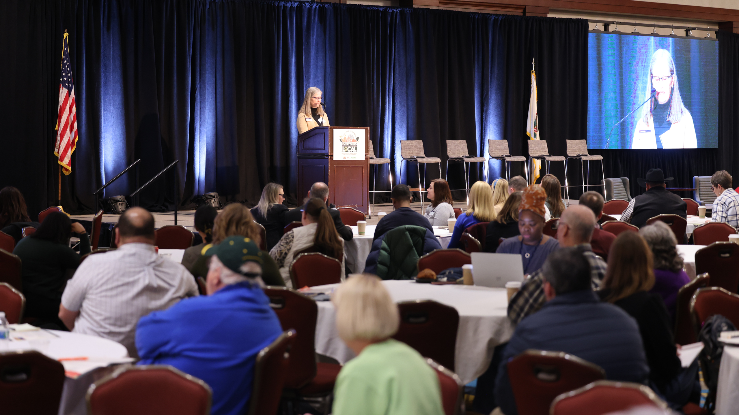 Woman speaking at a conference to a group of people sitting at tables.  